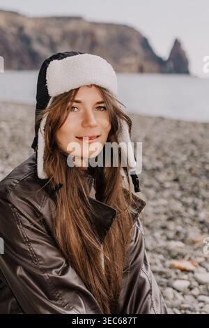 Donna Ushanka Beach bella giovane donna che indossa un caldo cappello invernale e una giacca in pelle su una costa di ciottoli con montagne Foto Stock
