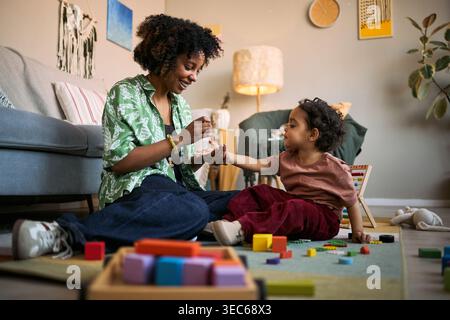 Madre afroamericana che applica l'igienizzatore alle mani del bambino mentre è seduta sul pavimento durante il gioco a casa Foto Stock