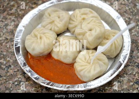 Momos tradizionali appena cotti serviti con chutney rosso piccante su un piatto di lamina monouso Foto Stock