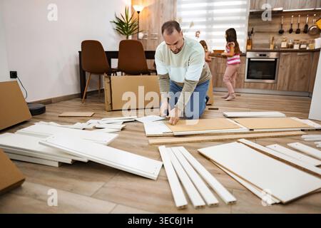 Un padre si concentra sull'assemblaggio di mobili flat pack in un soggiorno caldo e invitante, mentre due bambini giocano nelle vicinanze, creando un'atmosfera allegra durante il progetto. Foto Stock