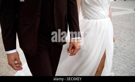 Elegante coppia in abito da sposa che si tiene per mano, cammina insieme. Sposo in abito scuro, sposa in abito bianco e fluente, momento intimo. Foto Stock