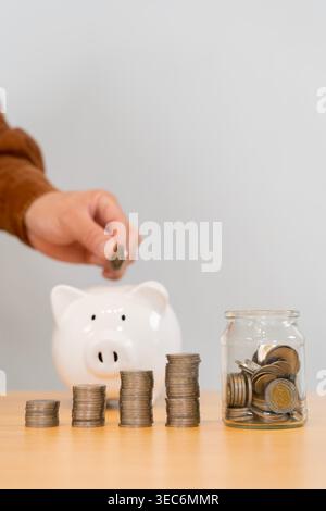 Mano per aggiungere una moneta a un salvadanaio dietro l'aumento delle pile di monete e un vaso di risparmio, che mostra risparmi costanti, disciplina di budget, crescita degli investimenti e pe Foto Stock