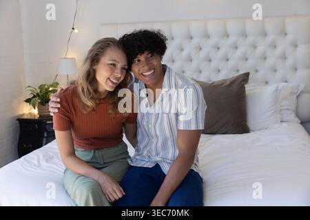 Coppia seduta sul letto appoggiata l'una verso l'altra, sorridendo vicino alla testiera capovolta e alle luci a corde Foto Stock