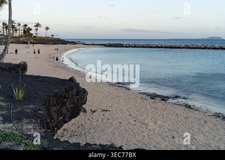 Una tranquilla spiaggia di Lanzarote, caratterizzata da sabbia soffice, onde dolci e una barriera di pietra. La gente passeggia lungo la riva, godendosi la serata. Foto Stock
