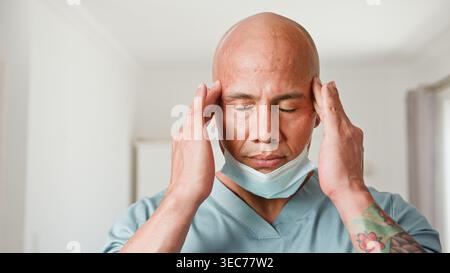 Medico di assistenza sanitaria stressato che indossa scrub e maschera con templi di tenuta del mal di testa Foto Stock