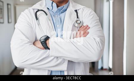 Medico maschio sicuro di sé con le braccia incrociate indossando cappotto bianco e stetoscopio in clinica Foto Stock