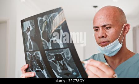 Medico che esamina la pellicola di scansione TC sinusale in clinica indossando maschera chirurgica e scrub Foto Stock