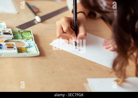 Una donna si concentra sulla pittura di lettere delicate con un pennello sottile su carta bianca. Lavora su un tavolo di legno pieno di oggetti d'arte e soffice l naturale Foto Stock