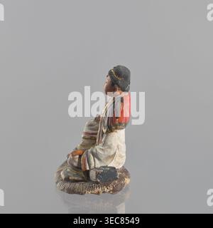Figurina di un uomo seduto, arte cinese, ceramica, abbigliamento maschile, figura seduta, statuetta maschile, XIX secolo, Cina, scultura, figurina, altezza 15 cm, Una piccola figurina dipinta di una persona seduta, mostrata in profilo su una base circolare con trama, caratterizzata da colori tra cui grigio, rosso, nero, bianco, marrone, beige, giallo e un tocco di verde; la figura indossa una testa nera ricoperta da accenti dorati, un indumento rosso sulle spalle e un abito per lo più bianco con finiture colorate, con vernice visibile usurata e scheggiata e alcune aree di agenti atmosferici più scuri sulla base e sull'abbigliamento. Foto Stock