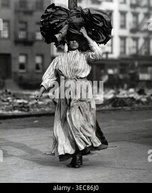 Questa fotografia del 1910 di Lewis Hine cattura una donna che trasporta un grosso fascio sulla testa lungo la Bowery di New York. La scena riflette le lotte quotidiane e il lavoro degli individui della classe operaia in ambienti urbani durante l'inizio del XX secolo. Foto Stock