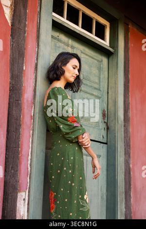 Una giovane donna con i capelli corti sta accanto a una porta verde su una vecchia casa rossa. Sembra premurosa e rilassata mentre si appoggia contro la porta in un semplice o Foto Stock
