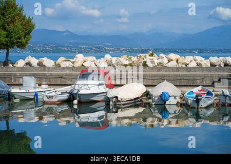 Fila di piccole barche colorate che si riflettono nell'acqua naturale del porticciolo sul lago, una giornata di sole con montagne e città sullo sfondo, destinazione del viaggio. Foto Stock