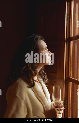Una giovane donna in una veste sta accanto a una finestra che regge un bicchiere di spumante. Guarda fuori, persa nel pensiero, mentre la luce del mattino riempie la stanza. La sce Foto Stock