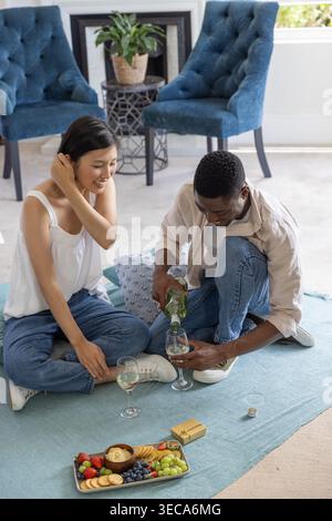 Coppia donna coreana, uomo afroamericano seduto su un tappeto a casa, che versa vino da una bottiglia verde Foto Stock