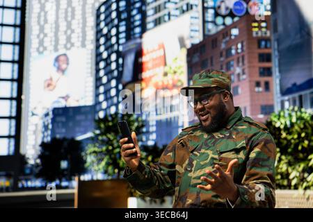 Soldato afroamericano allegro in uniforme che scorre su smartphone e tifa, ricevendo buone notizie di successo durante la pattuglia del centro città. Soldato che cammina lungo la trafficata strada del centro città. Foto Stock