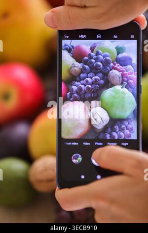 Primo piano verticale dello schermo di uno smartphone che cattura la natura morta di uva, pere e noci. Le mani di una persona sono visibili tenendo di nuovo il dispositivo Foto Stock
