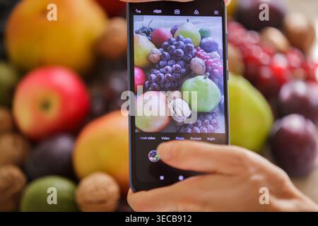 Primo piano di una persona che tiene uno smartphone e scatta una foto di una varietà di frutta biologica, tra cui uva, pere, mele e noci. Concentrati su t Foto Stock