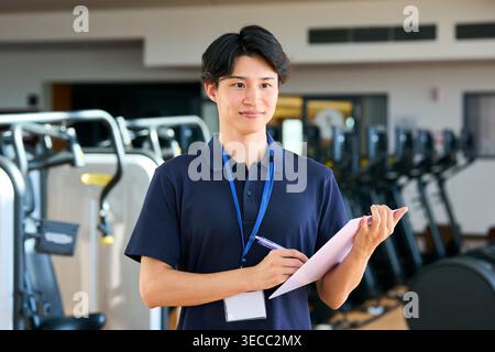 Un giovane personal trainer giapponese sorridente che scrive su documenti in una palestra Foto Stock