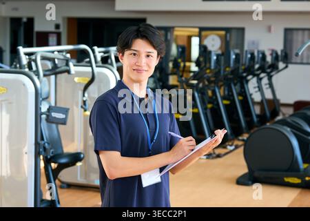 Sorridente giovane istruttore di palestra giapponese maschile che prende appunti con un appunti Foto Stock