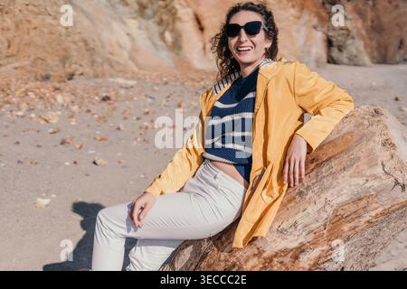 Woman Beach Smiling siede su un grande tronco di legno da mare vicino a una scogliera rocciosa sotto il sole Foto Stock