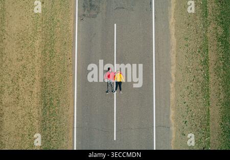 Vista aerea di una coppia di viaggiatori riposano su una strada vuota rettilinea, godendo di tranquillità con le mani dietro la testa Foto Stock