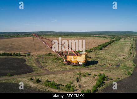 L'enorme escavatore dragline industriale poggia in un campo aperto, circondato da vegetazione sparsa e strade sterrate Foto Stock