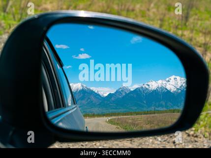 Attraversando maestose montagne innevate, un cielo azzurro si riflette nello specchio laterale dell'auto in una giornata intensa Foto Stock