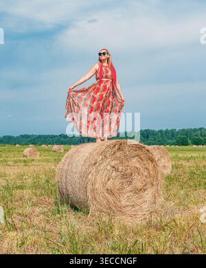 Donna in piedi su una grande balla di fieno in un campo estivo, che indossa un vestito che scorre e occhiali da sole sotto un cielo luminoso Foto Stock