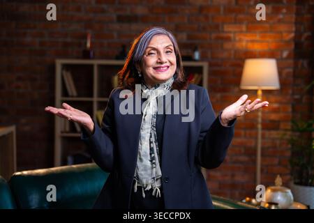 Donna d'affari indiana asiatica anziana in abbigliamento aziendale che lavora su un computer portatile mentre si siede su un comodo divano in un moderno ufficio sorride alla macchina fotografica e mostra Foto Stock