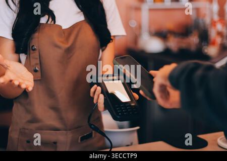 Barista asiatica anziana che riceve pagamenti mobili da un cliente tramite smartphone presso un bar che dimostra una moderna transazione senza contanti in un ambiente accogliente Foto Stock