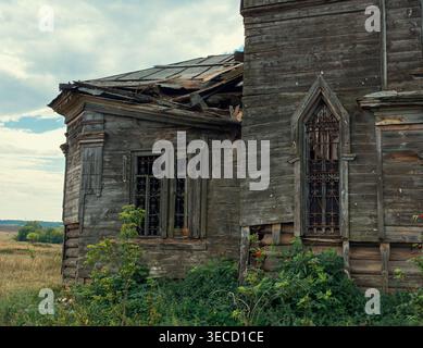 Edificio in legno fatiscente con tetto crollato e finestre gotiche ornate in un ampio campo rurale Foto Stock