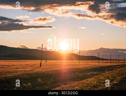 La luce dorata illumina un ampio campo di Altai mentre il sole tramonta dietro montagne lontane, con pali di servizio che fiancheggiano l'orizzonte Foto Stock
