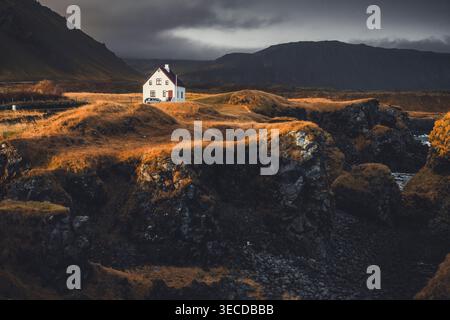 Una casa solitaria ad Arnarstapi si erge contro i paesaggi costieri selvaggi dell'Islanda, dove scogliere, oceano e silenzio si incontrano sotto vasti cieli. Foto Stock