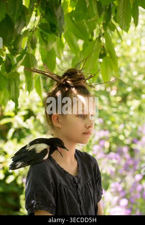 Una ragazza seria con un uccello magpie che le chiama sulla spalla in un giardino luminoso. Concetto di voce, espressione, voce e comunicazione. Foto Stock