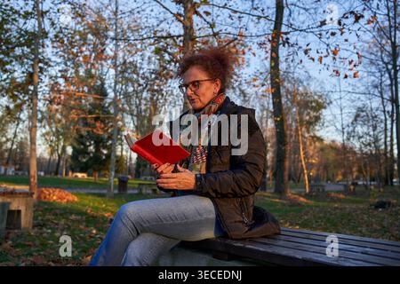 Donna di mezza età con capelli ricci rossi e occhiali seduti su una panchina del parco a leggere un libro coperto di rosso, concentrata e assorbita nella sua lettura in caldo autu Foto Stock