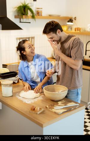 In una cucina luminosa, una donna coreana e un uomo caucasico condividono le risate mentre preparano. Ogni interazione gioiosa mostra la loro connessione attraverso il cibo. Foto Stock