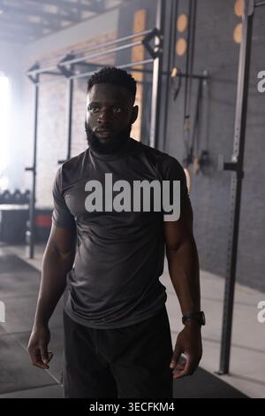 Uomo afro-americano in palestra con attrezzature e manubri, con camicia scura e pantaloncini neri. Forza, allenamento, pesi, barbell, rack, lighti Foto Stock