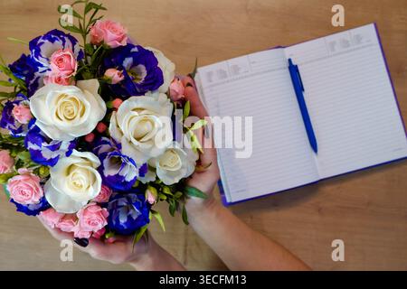 Le mani femminili tengono in mano un fiore di rosa. Quaderno, diario con penna su un tavolo di legno vicino a un mazzo di rose multicolore. Foto Stock