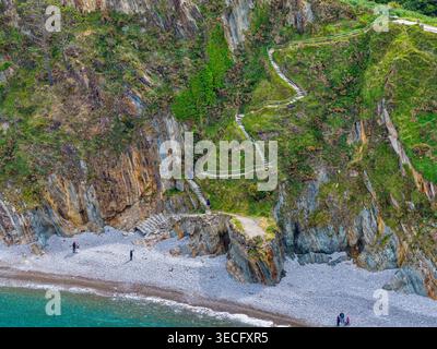 Veduta aerea del punto panoramico di Silence Beach con una tortuosa scalinata in pietra su ripide scogliere e persone che camminano giù per la spiaggia di ciottoli, Cudillero e le Asturie Foto Stock
