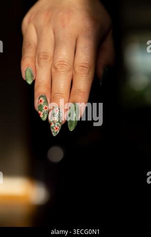 Una mano con lunghe unghie mostra un design verde lucido e floreale. L'impostazione è in interni con luce soffusa. L'attenzione è rivolta ai dettagli della nail art AN Foto Stock