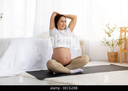 Donna attiva in gravidanza seduta su un tappetino da yoga che si allunga le braccia sopra la testa con un sorriso Foto Stock