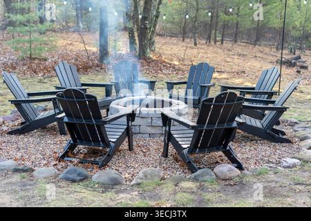 Area con posti a sedere all'aperto con sedie Adirondack intorno al caminetto nel cortile della foresta, atmosfera accogliente con luci a corda e fumo. Foto Stock
