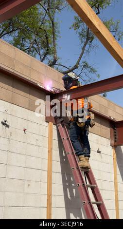 Il lavoratore edile salda un telaio metallico all'aperto con scintille e dispositivi di protezione presso un cantiere di Los Angeles Foto Stock