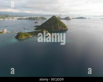 Vista aerea delle lussureggianti isole tropicali verdi e delle barche ancorate nelle acque turchesi della baia vicino all'isola di Flores sotto un morbido cielo nuvoloso a Labuan Foto Stock