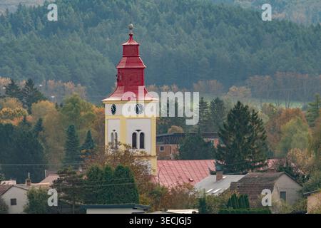 Vista aerea della torre della chiesa della Santissima Trinità con il suo tetto rosso e la facciata dell'orologio circondata da alberi autunnali e case del villaggio di Mosovce, Foto Stock