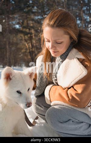 Una giovane donna si inginocchiò caldamente accanto a un adorabile cucciolo Akita bianco sulla neve. La luce del sole filtra tra tra gli alberi in un ambiente invernale sereno. Foto Stock