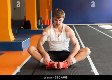 Un giovane sportivo che indossa una canotta bianca e pantaloncini neri si siede su un tappetino da yoga in una palestra moderna, concentrandosi su esercizi di flessibilità per migliorare l'atletica Foto Stock