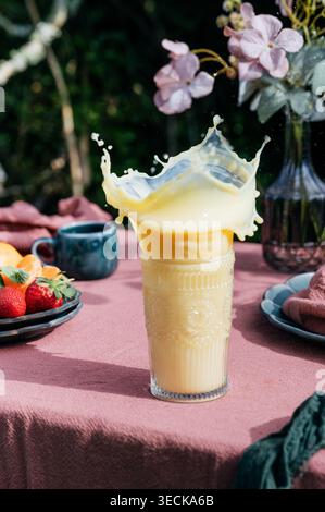Succo di arancia, ananas e albicocca che spruzza in un bicchiere alto su un tavolo da colazione con frutta fresca, fiori e luce soffusa del mattino, creando un vibran Foto Stock