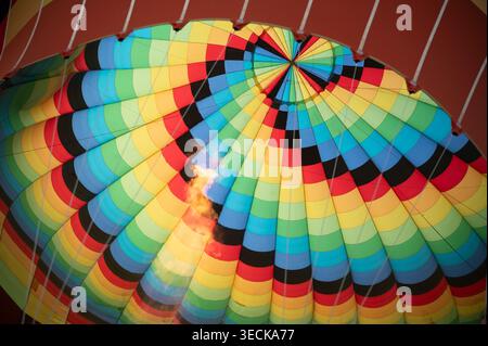 Vista ravvicinata all'interno di una colorata cappottatura di una mongolfiera durante il volo, che mostra vivaci pannelli di tessuto, linee di sospensione e una fiamma di riscaldamento luminosa Foto Stock