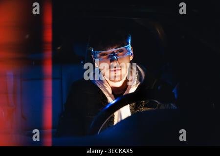 Ritratto ravvicinato di un giovane uomo che indossa una visiera olografica luminosa di notte, circondato da riflessi al neon scuro, che evoca la cybersicurezza, l'hacking, un Foto Stock